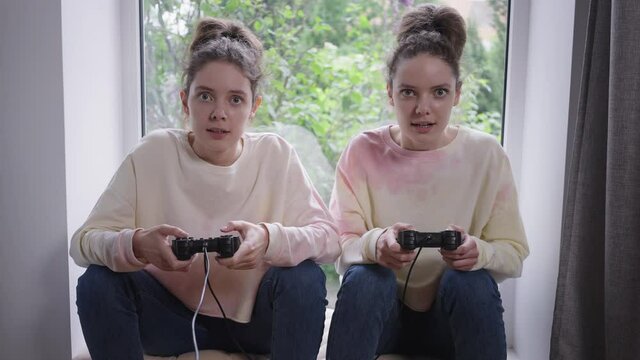 Front view absorbed twin gamers playing with joysticks sitting on windowsill at home indoors looking at camera. Portrait of cheerful beautiful Caucasian identical women gaming having fun