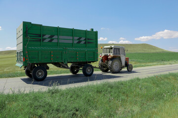 Green tractor with trailer is going on the village road.  It has huge wheels. Roadside is green...