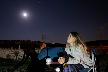 A girl camping in the moonlight at night. Teapot behind her. She is watching the sky.