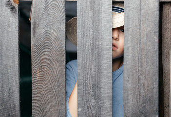 Five- to six-year-old kid in a straw hat peeps through a wooden village fence. Copy space