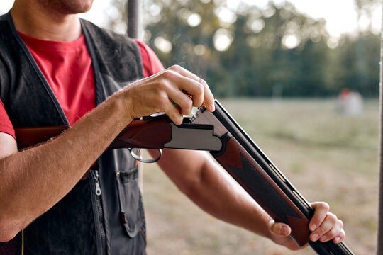 Cropped young man in uniform charges pump-action shotgun in an outdoor range. Skilled experienced man is preparing to shoot. Firearms for sports shooting, hobby. Copy space. close-up hands