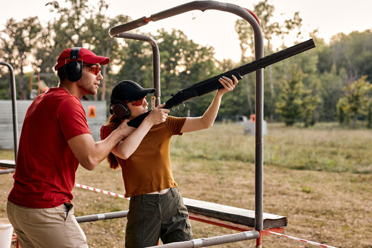 young man and woman in goggles and headset preparing to shoot, ready to shoot. Skilled experienced man is teaching female in outdoor range. Firearms for sports shooting, hobby. Side view.