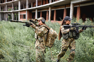 Military ladies women in tactical gear posing for photo in field at summer season. Female is Wearing green camo uniform and weapon with suppressor, abandoned building in the background