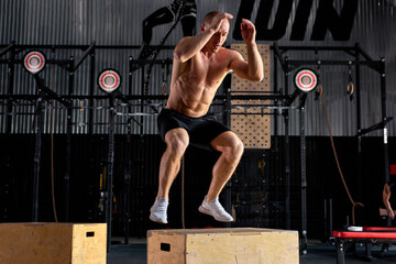 Young caucasian shirtless male is jumping on cross fit wooden box, at modern sportive gym. Athlete young guy in good perfect physical shape is engaged in sport, fitness. side view. alone.