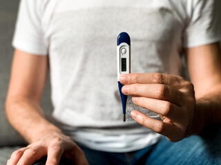 Fototapeta premium Man is checking temperature with digital thermometer. A man sitting on gray sofa with thermometer in hands. Holding thermometer, checking temperature. Seasonal health problems - flu, angina, fever