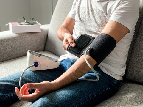 Man Is Checking Blood Pressure At Home. Sitting On A Gray Sofa. Seasonal Health Issues. Hypertension Or Hypertension Seasonal Disorder And Problems. Taking Care Of Health, Looking After Yourself