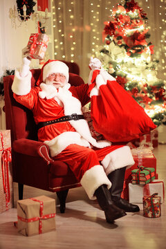 Santa Claus with a real white beard in a traditional red and white suit and round glasses is smiling sitting on a chair and holding a bag of gifts