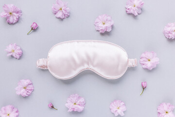Pink satin eye sleeping mask and fresh spring Japanese cherry blossom flowers on pastel background. Insomnia, sleeplessness disorder, beauty sleep concept. Close up, copy space, flat lay. Accessories