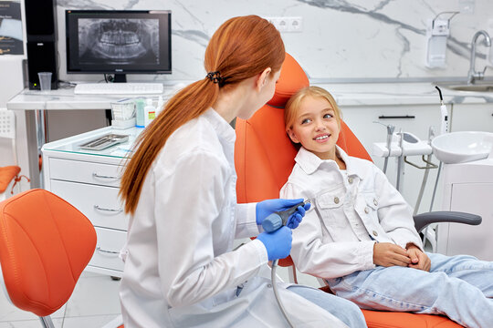 Dentist in gloves holding dental drill while treating child girl patient in modern dental office clinic, side view on adorbale excited child girl sitting on couch waiting for treatment. - Powered by Adobe
