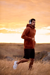 Handsome athletic male in hoodie training jogging outdoors, listening to music, on field, fit european sportsman in headphones, running alone. sport, fitness, healthy lifestyle concept.