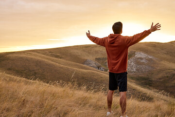 caucasian male stands against the backdrop of mountain landscape with hands raised high In the...