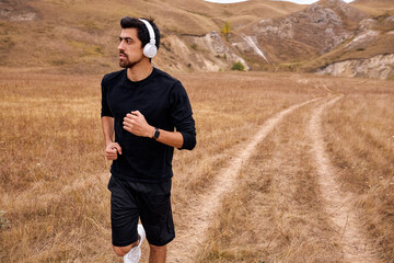 Fototapeta premium Bearded Man running,jogging, sprinting in mountains. Fit male fitness runner during outdoor workout. Young athlete caucasian man in black sportswear tracksuit and headphones listening to music.