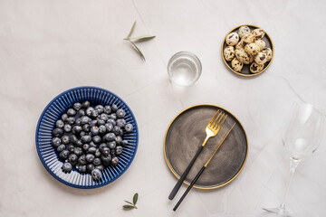 White marble table with blue plate filled with ripe blueberries, vintage plate with cutlery and another plate with quail eggs and olive leaves