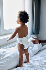 beautiful curious black dark skinned toddler girl stand on bed looking through the window,...