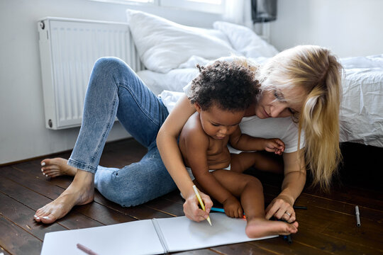 blonde female and small adorable daughter sitting on wooden warm floor at modern home drawing on paper album with colored pencils. Having fun, child development, family leisure activity hobby concept
