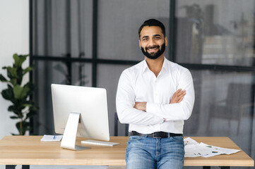 Portrait of a successful confident Indian man, mentor, manager or IT specialist, wearing a white shirt, standing near desk in the office with arms crossed, looking at the camera and friendly smiling