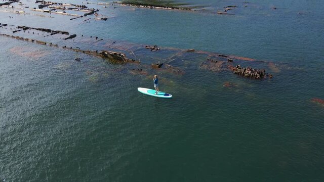 Aerial shot. Young women is having fun. Stand Up Paddling in the sea. SUP. Paddling among debrees of a sunk metal ships