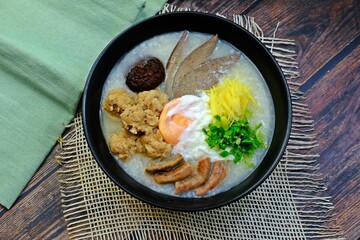 Congee or Porridge with minced pork, Shiitake Mushroom, soft-boiled egg, pork liver, pork intestines, ginger sliced, spring onion sliced, coriander sliced in black bowl on wooden table.