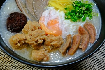 Congee or Porridge with minced pork, Shiitake Mushroom, soft-boiled egg, pork liver, pork intestines, ginger sliced, spring onion sliced, coriander sliced in black bowl on wooden table.