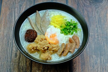 Congee or Porridge with minced pork, Shiitake Mushroom, soft-boiled egg, pork liver, pork intestines, ginger sliced, spring onion sliced, coriander sliced in black bowl on wooden table.