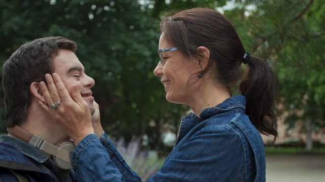 Happy mature mother kissing on forehead her adult son with Down syndrome outdoors in town.