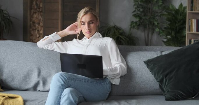 Thoughtful female blogger using laptop for writing posts in social network. Professional busy freelancer sitting on coach and search for inspiration and solving problems. Young woman chatting
