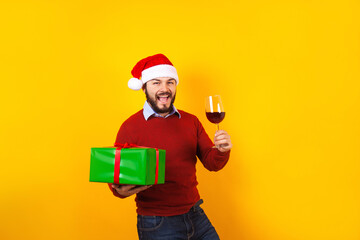 young latin man holding a glass of wine with santa hat and christmas presents on a yellow background in Mexico Latin America