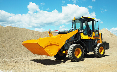 Obraz na płótnie Canvas Black-yellow front loader with small wheels against the background of a large pile of stone sand and a blue sky with white clouds.