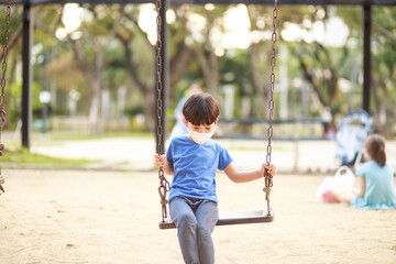 A cute Asian boy wearing a mask is playing on a swing in the playground during the daytime in summer. Outdoor activities. Play Makes Ideas Believe External education. portrait