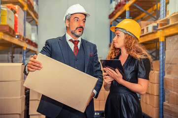 Manager holding cardboard box and talking to supervisor holding digital tablet with blurred background stock of  inventory and products ready for shipment.