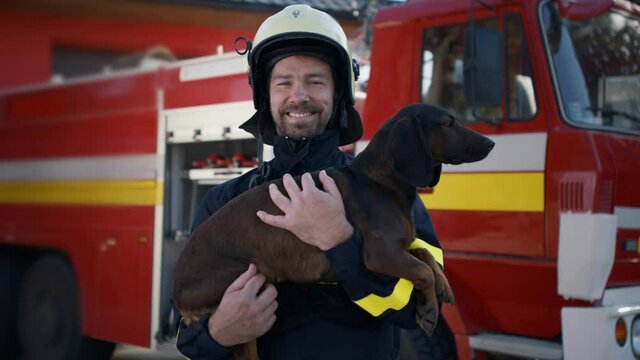 Happy mature firefighter man holding dog and looking at camera with fire truck in background