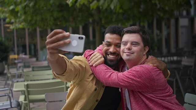 Young man with Down syndrome and his mentoring friend sitting and taking selfie outdoors in cafe