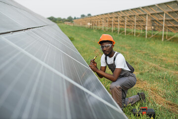 African american man in uniform and helmet using screwdriver for repairing solar panels on station. Installation of photovoltaic cells. Green energy concept.