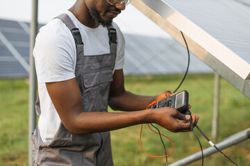Close up of african american man in grey overalls measuring resistance in solar panels outdoors. Competent technician using multimeter during inspection on station. - obrazy, fototapety, plakaty