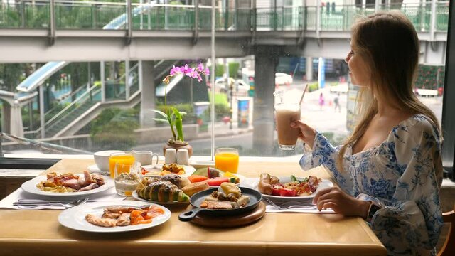 Modern female businesswoman eat breakfast buffet in urban cafe with city life, cars drive outside window. Young woman drink cold coffee in hotel restaurant during dinner or lunch. Slow motion.