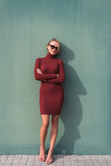 vertical portrait of an attractive blonde woman leaning on a green wall. She is dressing a garnet...