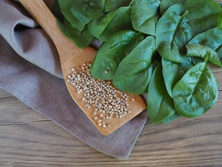 Seeds in a wooden spoon and fresh young spinach leaves with a napkin on a wooden table, flat layout. Spinacia leaf salad with vitamins for dietary nutrition. Farming and gardening - obrazy, fototapety, plakaty