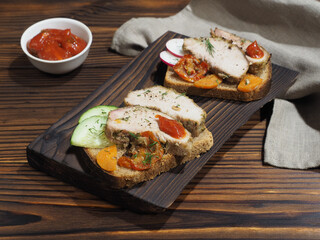 Toasts, bruschetta with slices of baked pork ham, grilled and fresh vegetables on rye bread, tomato sauce on a dark wooden table. Delicious snack, appetizer with natural meat and vegetables for lunch