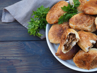 Fried pies with cabbage in a plate with a napkin, herb parsley on a dark wooden table, closeup, top view, flat layout, copy space. Traditional delicious hot snack pirozhki. Russian, Ukrainian cuisine