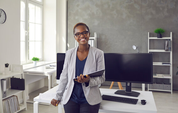 Portrait Of Happy Successful Satisfied Young Business Lady At Work. Beautiful Black Woman With Short Hair Smiling And Looking At Camera Standing In Office Interior By Table With Desktop Computers