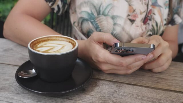 Girl with pink nails watches the news in a fashionable new phone, sitting at a wooden table on which there is coffee. Person puts likes on phone over a cup of morning coffee in cafeteria. Slow motion.
