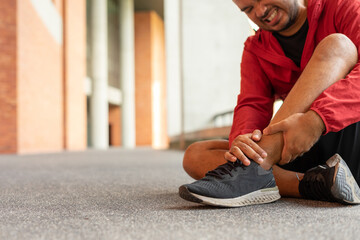 Closeup of sportsman wearing a red jacket injured his ankle during exercise outdoors brick wall on city street. Athletic male in hoodie sweatshirt red flipped ankle from joint pain in urban city park.