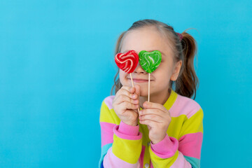 portrait of a cute little girl with lollipops in a striped jacket covering her eyes with them, a large candy on a stick. The concept of sweets and confectionery. Blue background, space for text