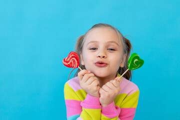 portrait of a joyful little girl with lollipops in a striped jacket, a large candy on a stick. The concept of sweets and confectionery. Blue background, photo studio, place for text