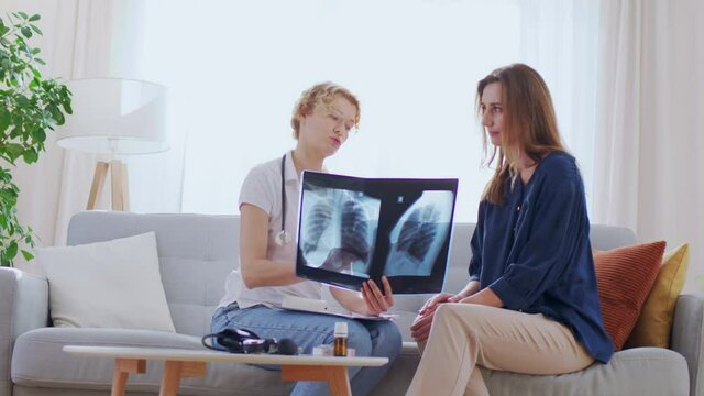 Attractive and smiling woman called young blonde hair nurse wearing in special white coat at home. They discuss x-ray picture while sitting on comfort couch in light colors interior