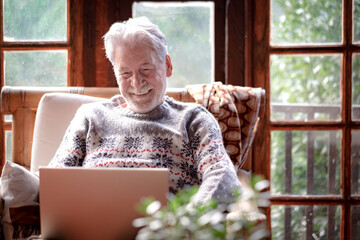 Smiling senior man in winter sweater sitting in living room using laptop computer. Carefree elderly...