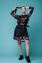 High fashion photo of a beautiful elegant young woman in a pretty black dress with colored patterns, sneakers posing over turquoise, blue background. Studio Shot. 