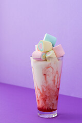 Glass cup with milkshake and fruit syrup, multicolored marshmallows on bright purple background, holiday or birthday concept