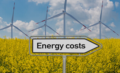 Energy costs word on a direction sign in front of a rapeseed field and wind farm, 