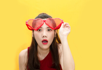 Asian beautiful woman with red theme on yellow background are wearing red big heart shape glasses looking to something with shocking and surprising and unbelievable.